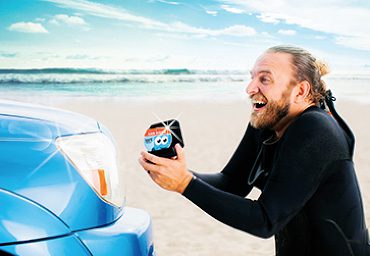 A man on the beach in a wetsuit proposing to his car with a Soapy Joe's air freshener.