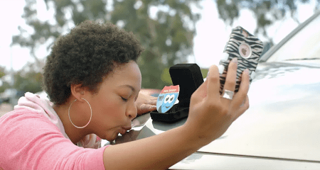 A woman taking a selfie of her kissing her car while she holds a Soapy Joe's air freshener in a ring box.