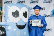 Graduate in cap and gown with diploma standing next to Soapy Joe mascot